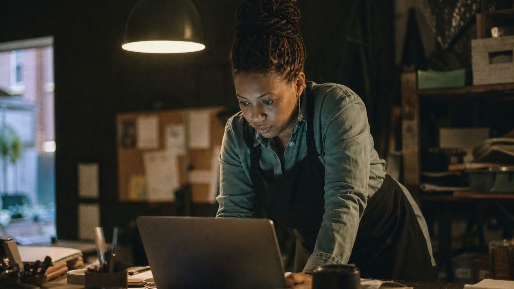 A small-business owner in an apron leaning over a laptop on a cluttered back-office desk late in the day, photographed in Durham