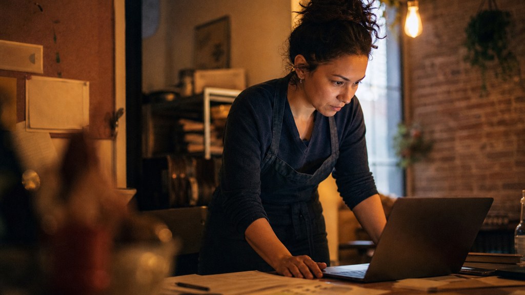 A small-business owner in an apron leaning over a laptop on a cluttered back-office desk late in the day, photographed in Durham