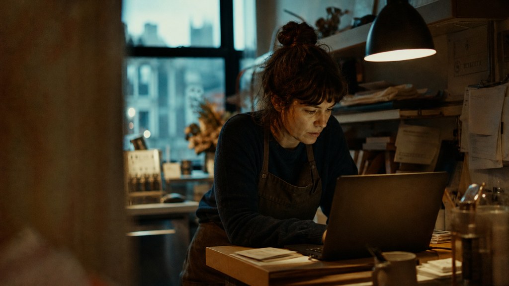 A small-business owner in an apron leaning over a laptop on a cluttered back-office desk late in the day, photographed in Durham