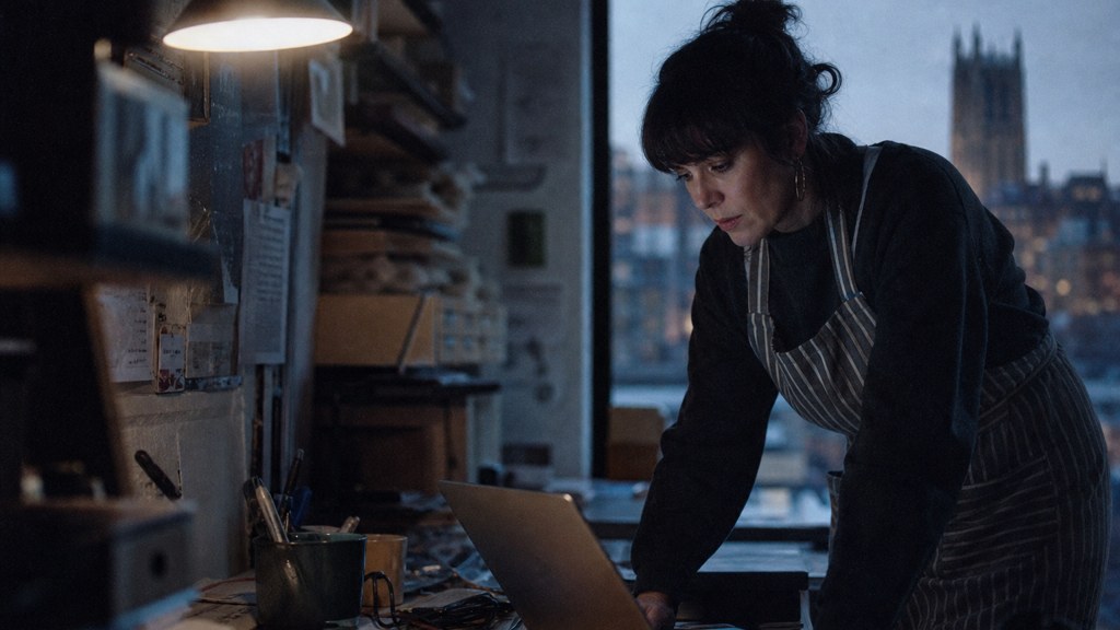 A small-business owner in an apron leaning over a laptop on a cluttered back-office desk late in the day, photographed in Durham