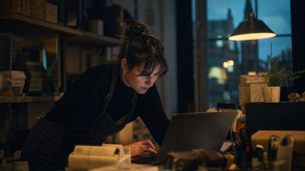 A small-business owner in an apron leaning over a laptop on a cluttered back-office desk late in the day, photographed in Edinburgh