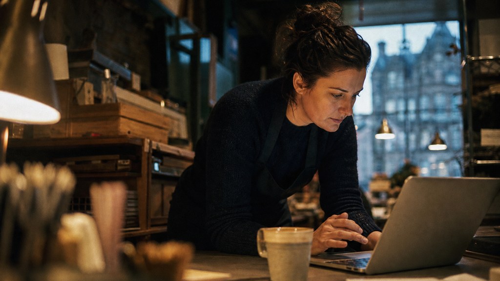A small-business owner in an apron leaning over a laptop on a cluttered back-office desk late in the day, photographed in Edinburgh