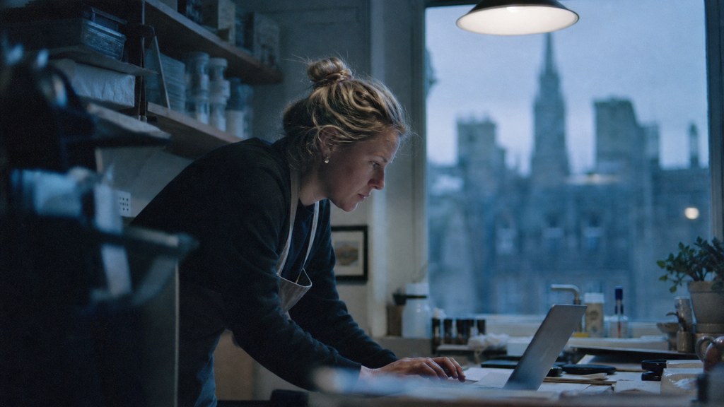 A small-business owner in an apron leaning over a laptop on a cluttered back-office desk late in the day, photographed in Edinburgh