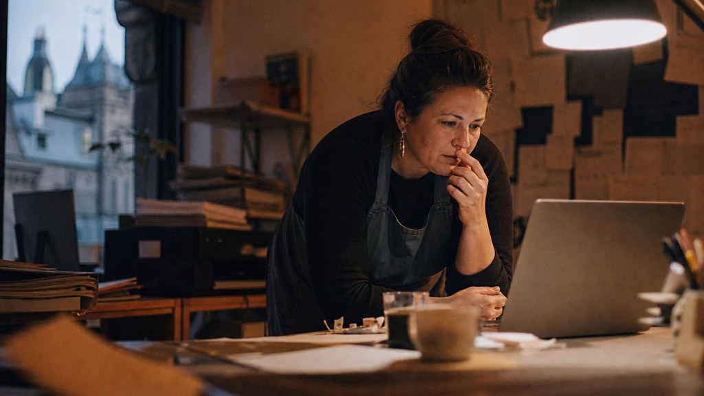 A small-business owner in an apron leaning over a laptop on a cluttered back-office desk late in the day, photographed in Edinburgh