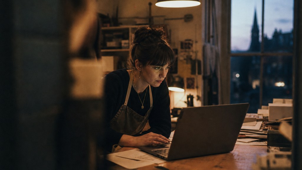 A small-business owner in an apron leaning over a laptop on a cluttered back-office desk late in the day, photographed in Edinburgh