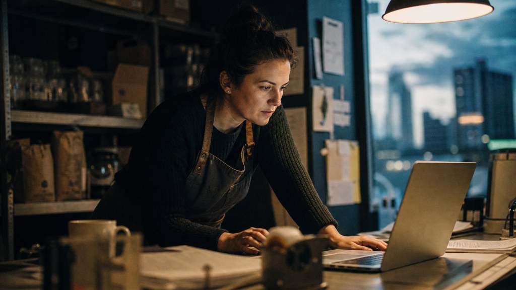 A small-business owner in an apron leaning over a laptop on a cluttered back-office desk late in the day, photographed in Edmonton