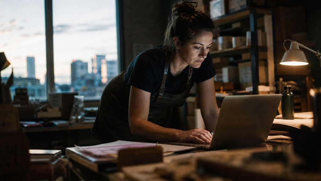 A small-business owner in an apron leaning over a laptop on a cluttered back-office desk late in the day, photographed in Edmonton