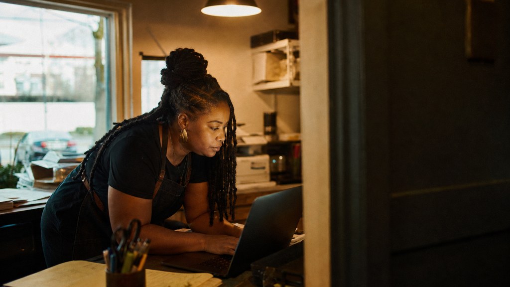 A small-business owner in an apron leaning over a laptop on a cluttered back-office desk late in the day, photographed in Fayetteville