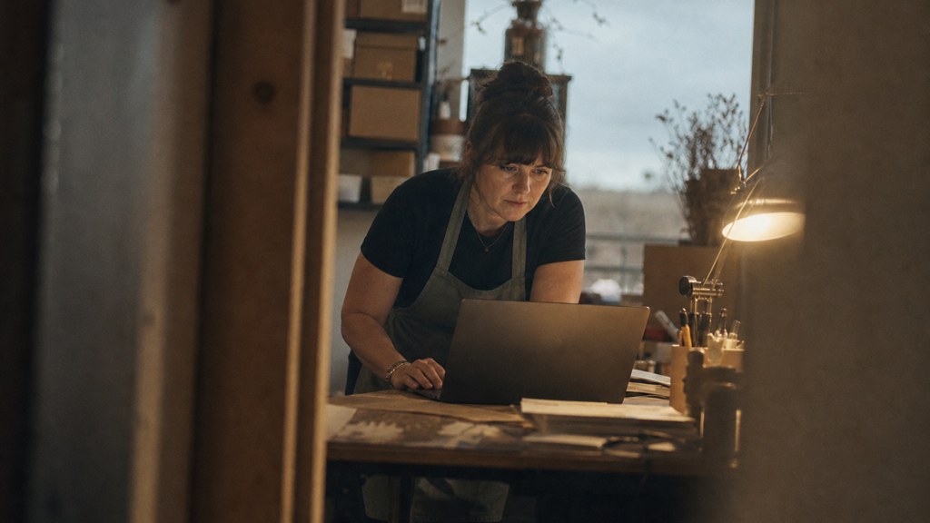 A small-business owner in an apron leaning over a laptop on a cluttered back-office desk late in the day, photographed in Fayetteville