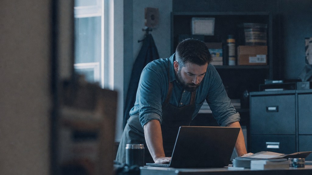 A small-business owner in an apron leaning over a laptop on a cluttered back-office desk late in the day, photographed in Fayetteville