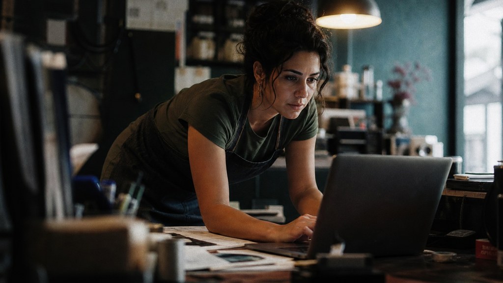 A small-business owner in an apron leaning over a laptop on a cluttered back-office desk late in the day, photographed in Fresno