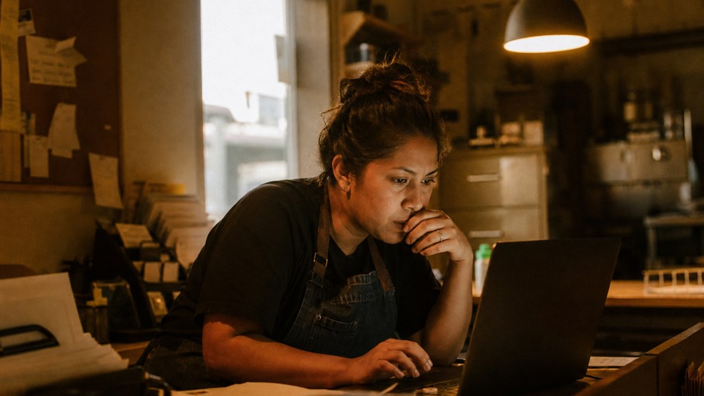 A small-business owner in an apron leaning over a laptop on a cluttered back-office desk late in the day, photographed in Fresno