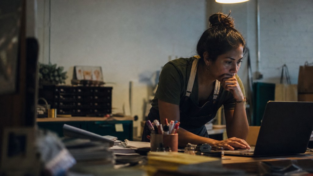 A small-business owner in an apron leaning over a laptop on a cluttered back-office desk late in the day, photographed in Fresno