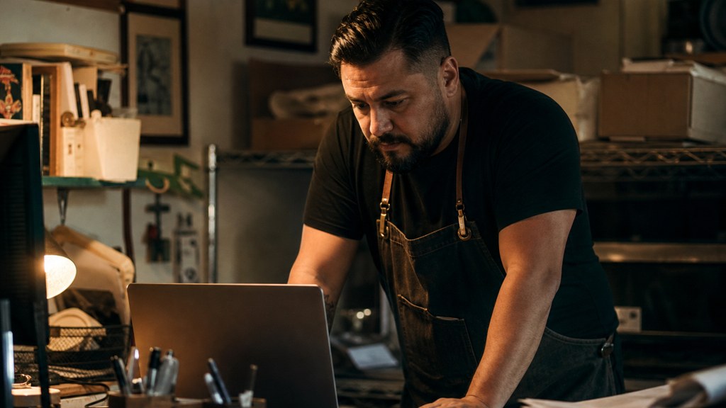A small-business owner in an apron leaning over a laptop on a cluttered back-office desk late in the day, photographed in Fresno