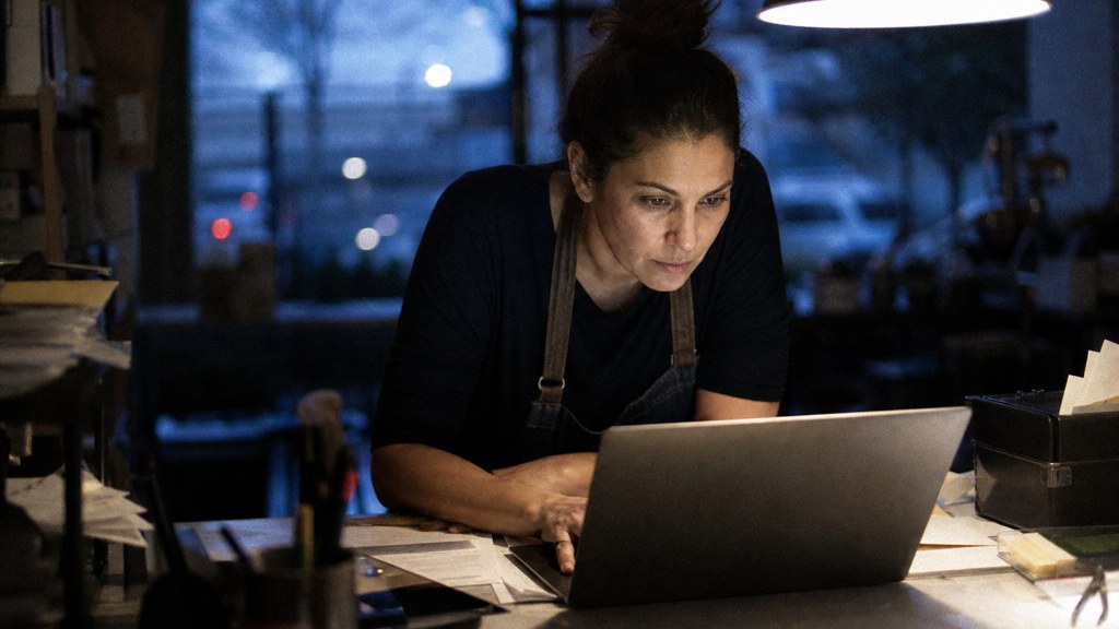 A small-business owner in an apron leaning over a laptop on a cluttered back-office desk late in the day, photographed in Frisco