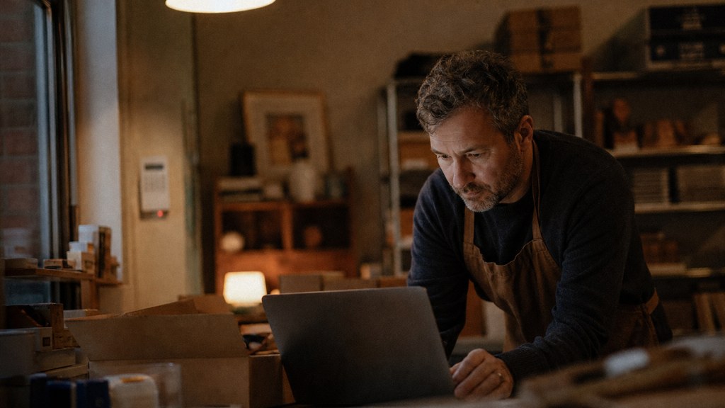 A small-business owner in an apron leaning over a laptop on a cluttered back-office desk late in the day, photographed in Frisco