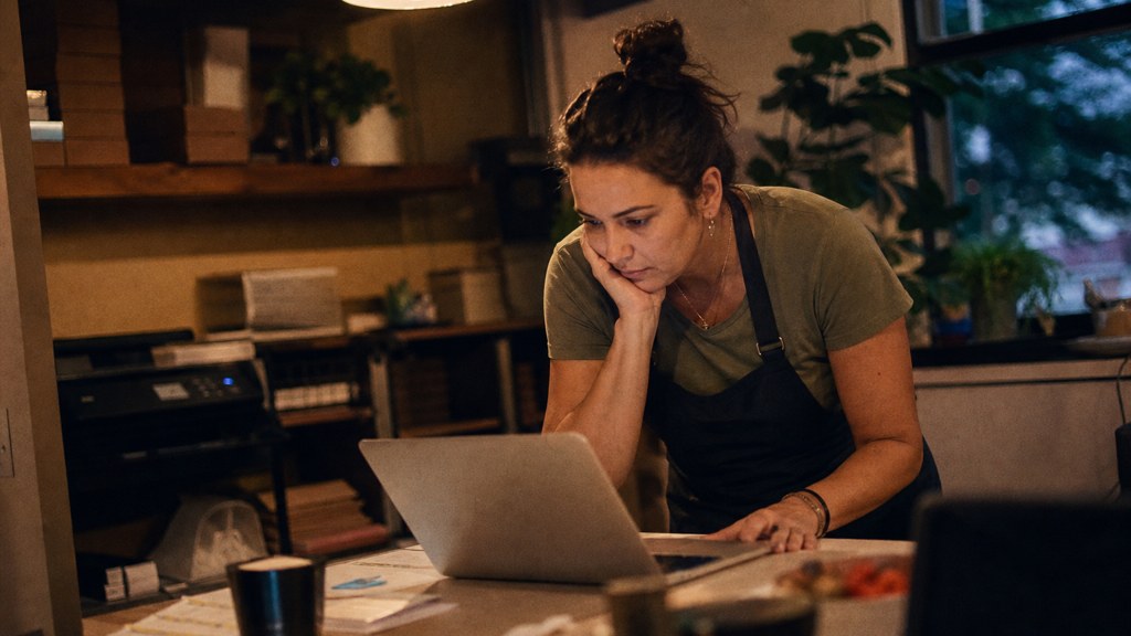 A small-business owner in an apron leaning over a laptop on a cluttered back-office desk late in the day, photographed in Frisco