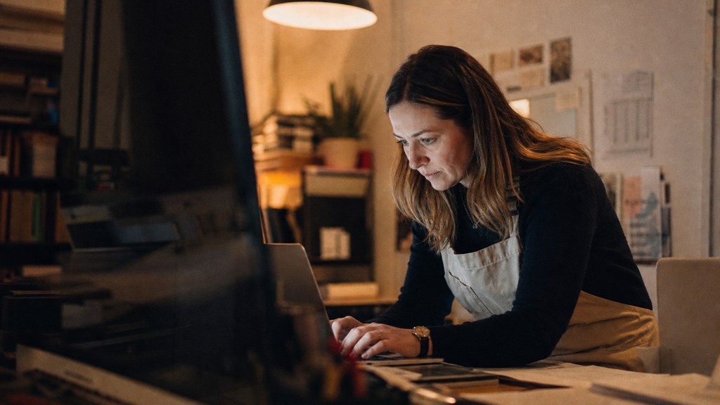 A small-business owner in an apron leaning over a laptop on a cluttered back-office desk late in the day, photographed in Frisco