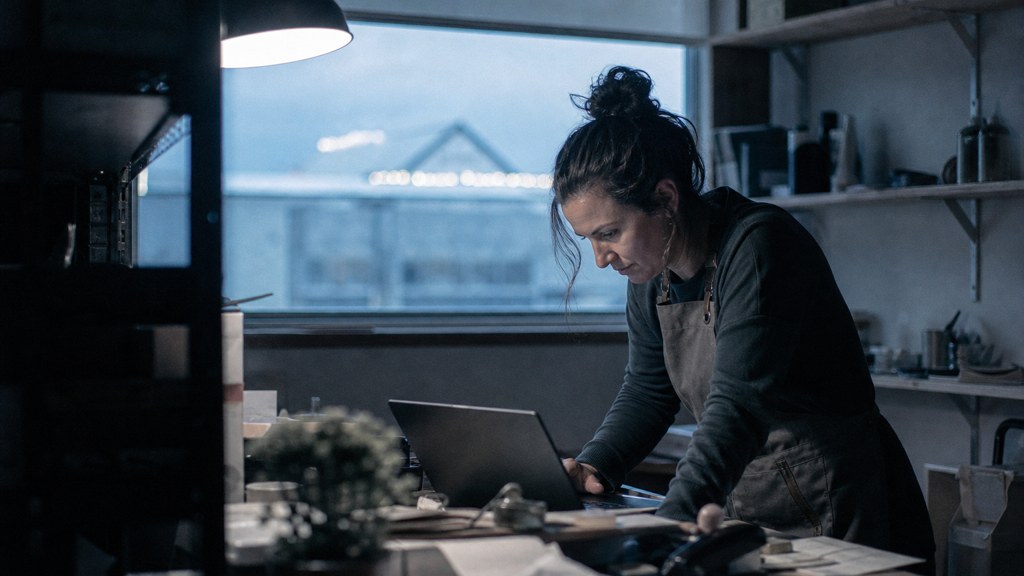 A small-business owner in an apron leaning over a laptop on a cluttered back-office desk late in the day, photographed in Frisco