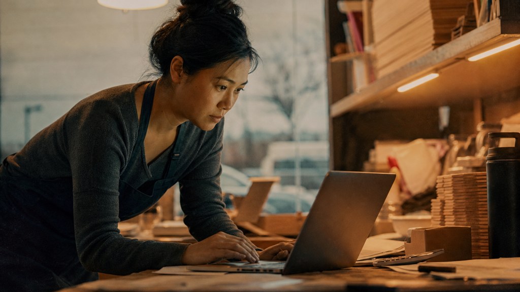 A small-business owner in an apron leaning over a laptop on a cluttered back-office desk late in the day, photographed in Frisco