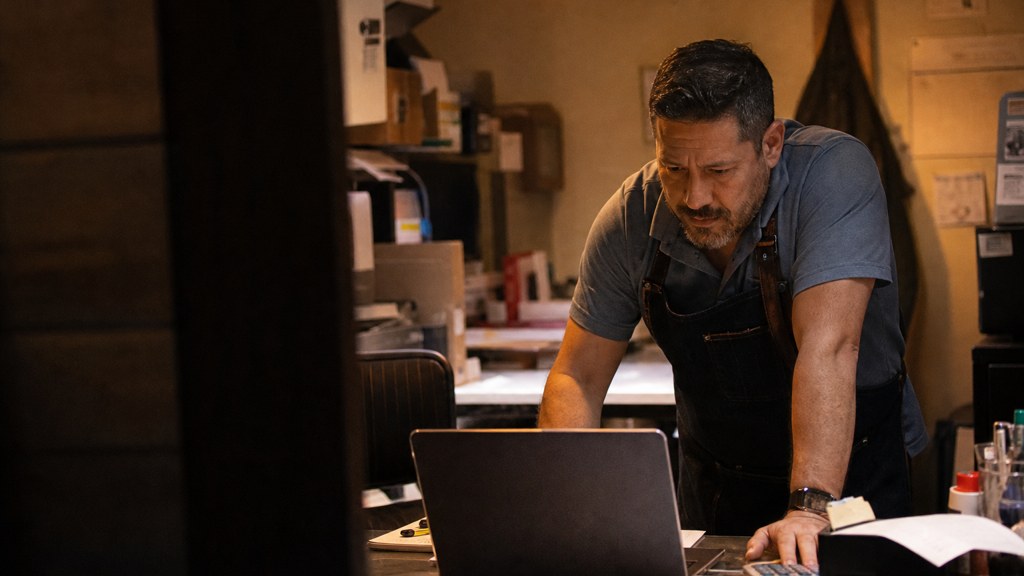 A small-business owner in an apron leaning over a laptop on a cluttered back-office desk late in the day, photographed in Garland