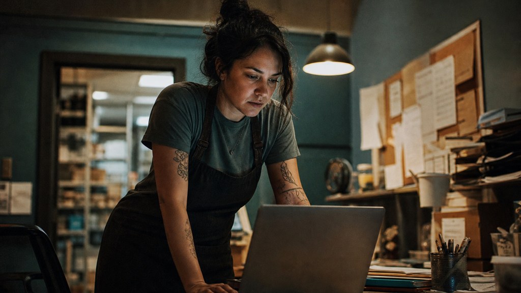 A small-business owner in an apron leaning over a laptop on a cluttered back-office desk late in the day, photographed in Garland