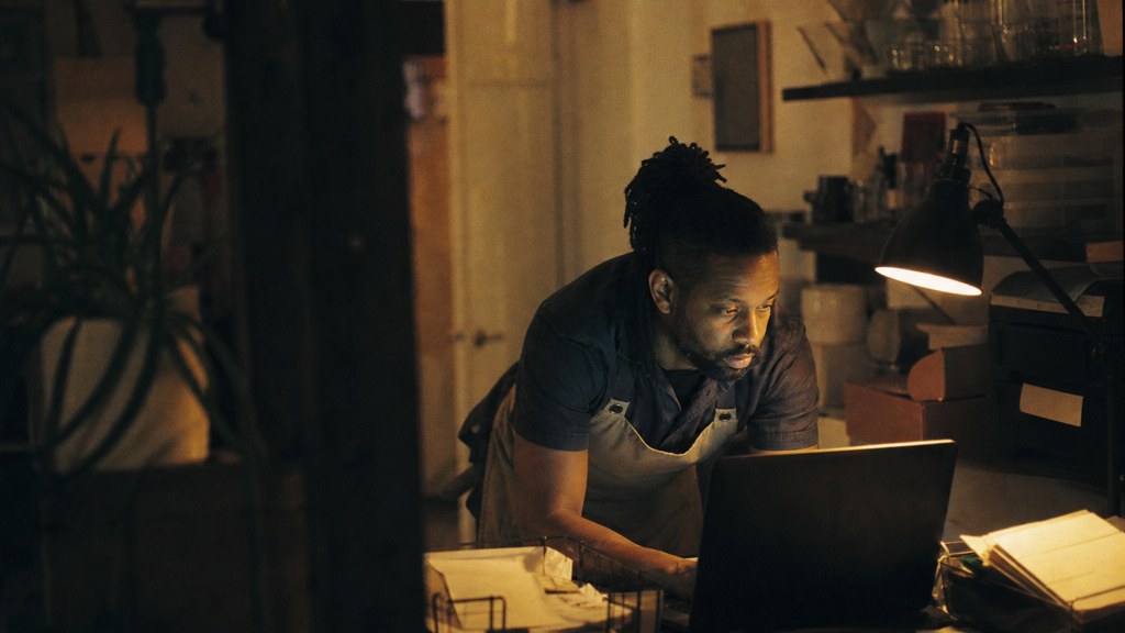 A small-business owner in an apron leaning over a laptop on a cluttered back-office desk late in the day, photographed in Garland