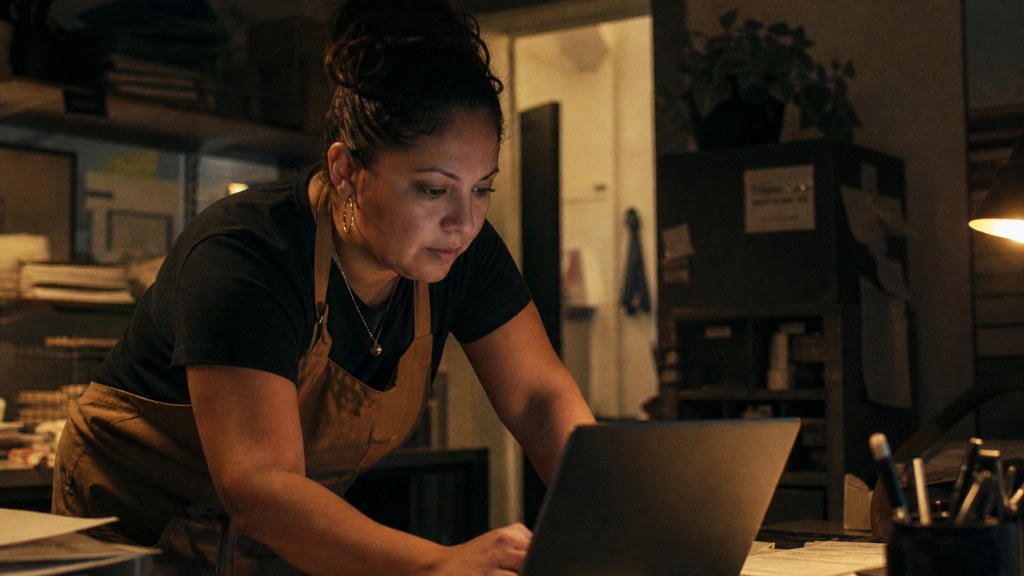 A small-business owner in an apron leaning over a laptop on a cluttered back-office desk late in the day, photographed in Garland
