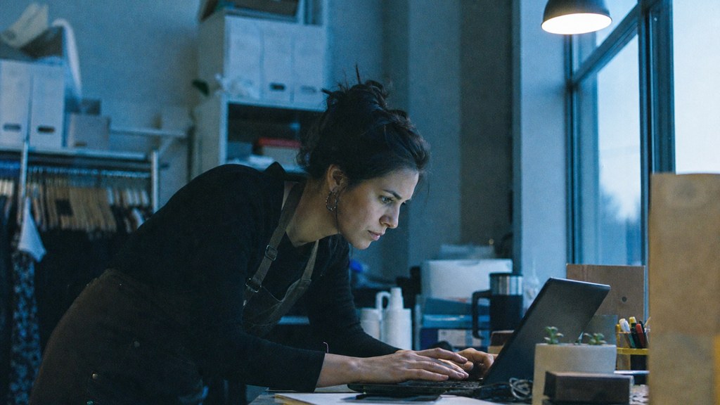 A small-business owner in an apron leaning over a laptop on a cluttered back-office desk late in the day, photographed in Garland