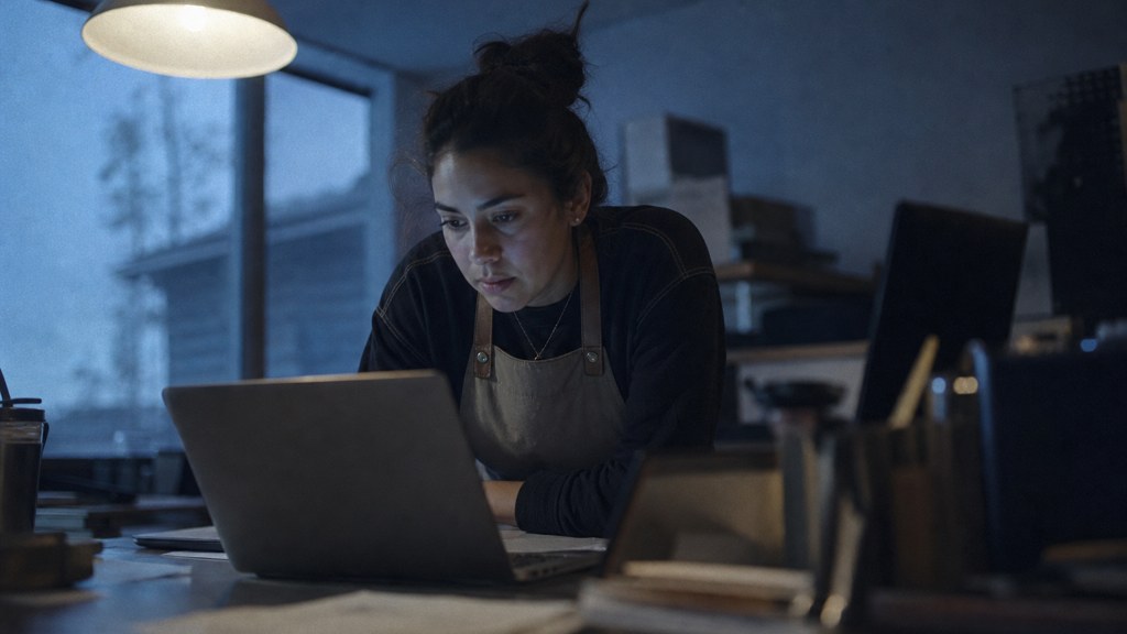 A small-business owner in an apron leaning over a laptop on a cluttered back-office desk late in the day, photographed in Garland