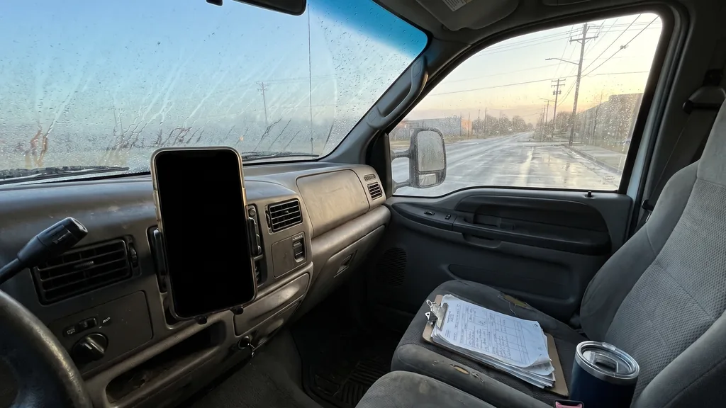 Editorial photograph of a restoration contractor truck cab at dawn with a phone mounted on the dash and a clipboard of job tickets on the passenger seat, no people, text or signage