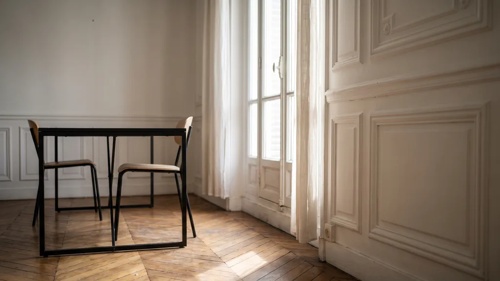 Editorial photograph of an empty workspace corner in a Parisian haussmannian office with herringbone parquet, white moulded panelling, a slim black metal table and two empty chairs by a tall french window, no people and no signage