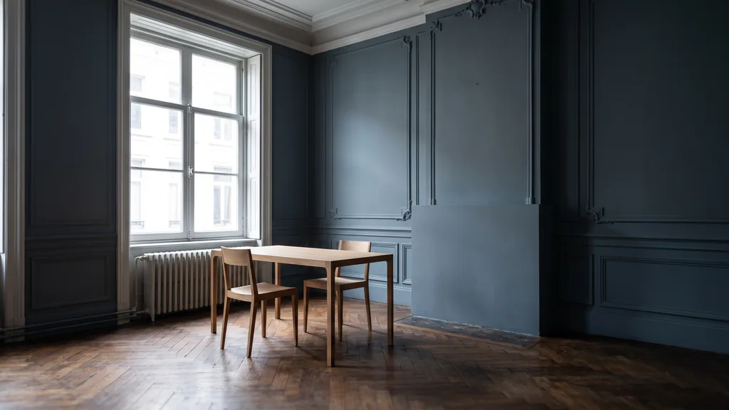 Editorial photograph of an empty workspace corner in a Brussels townhouse office with dark parquet, deep blue-grey walls, a slim oak table and two empty chairs by a tall sash window, no people and no signage