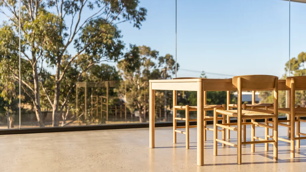 Editorial photograph of an empty meeting corner in a West Perth low-rise office with a light polished floor, a tall window over eucalyptus trees and blue sky, a plain pale table and empty chairs, no people and no signage