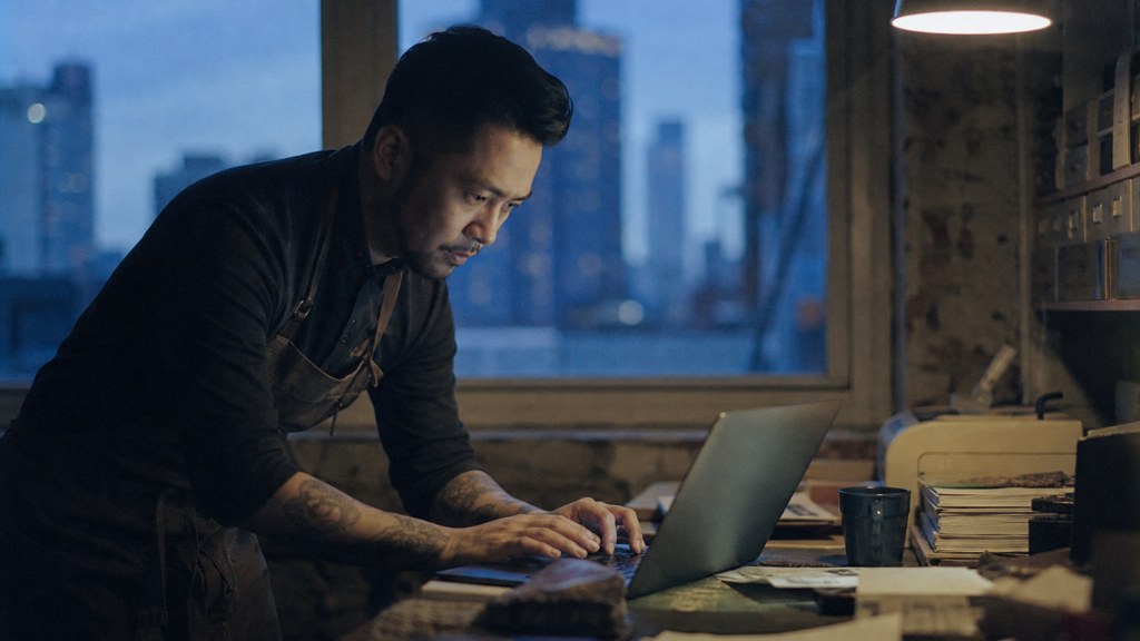 A small-business owner in an apron leaning over a laptop on a cluttered back-office desk late in the day, photographed in New York