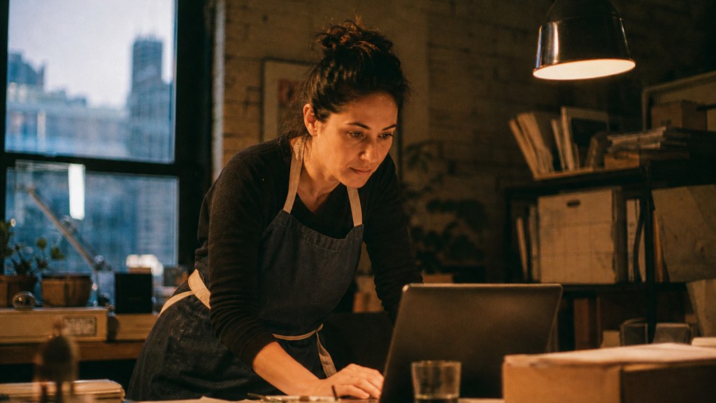 A small-business owner in an apron leaning over a laptop on a cluttered back-office desk late in the day, photographed in New York
