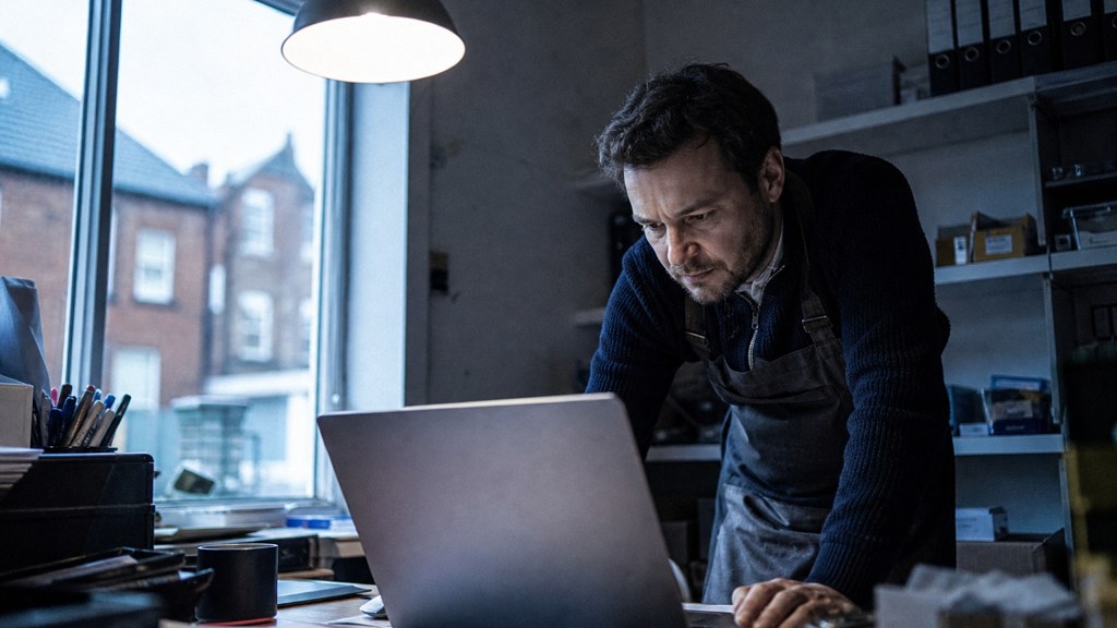 A small-business owner in an apron leaning over a laptop on a cluttered back-office desk late in the day, photographed in Norfolk