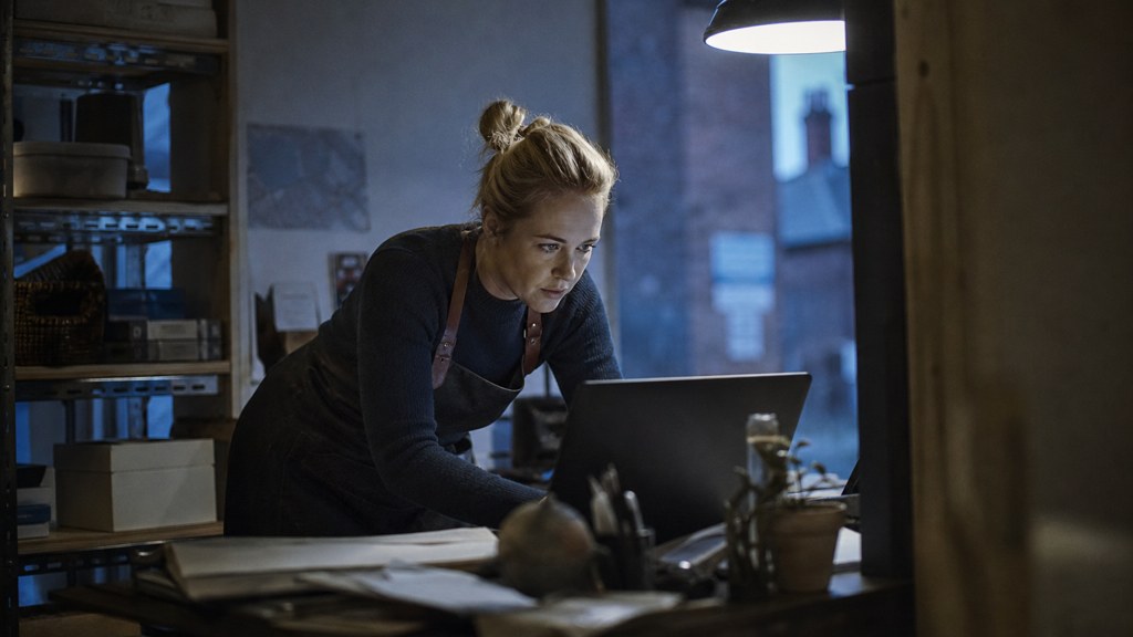 A small-business owner in an apron leaning over a laptop on a cluttered back-office desk late in the day, photographed in Norfolk