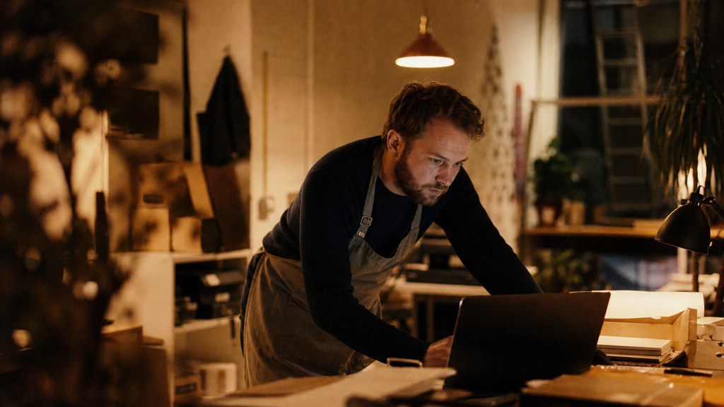 A small-business owner in an apron leaning over a laptop on a cluttered back-office desk late in the day, photographed in Norfolk