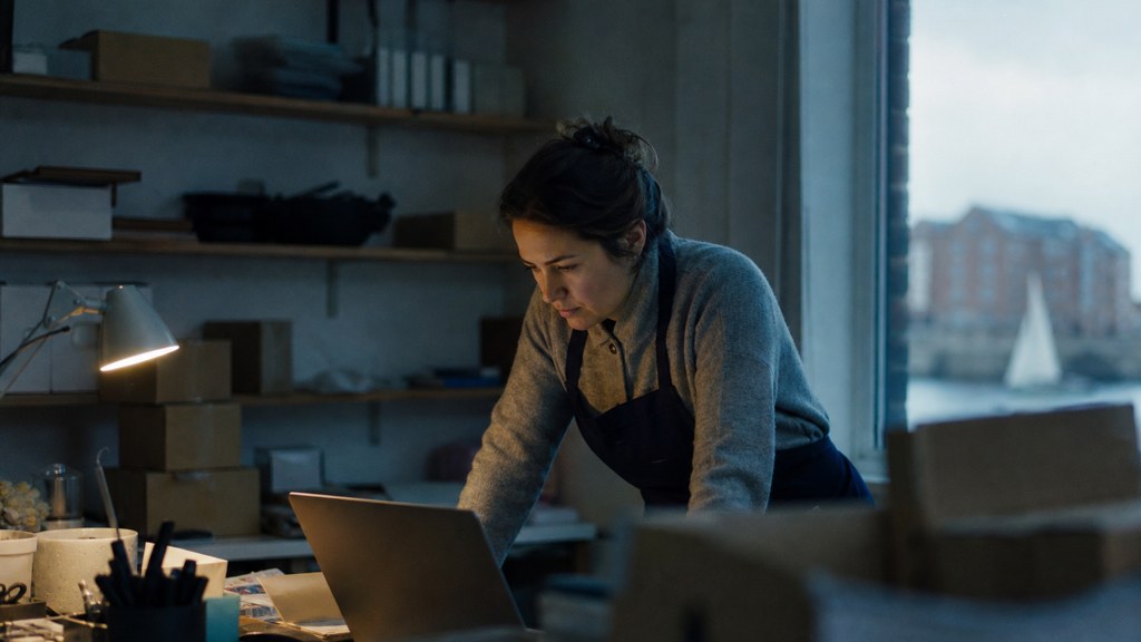 A small-business owner in an apron leaning over a laptop on a cluttered back-office desk late in the day, photographed in Norfolk