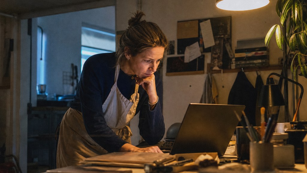 A small-business owner in an apron leaning over a laptop on a cluttered back-office desk late in the day, photographed in Norfolk