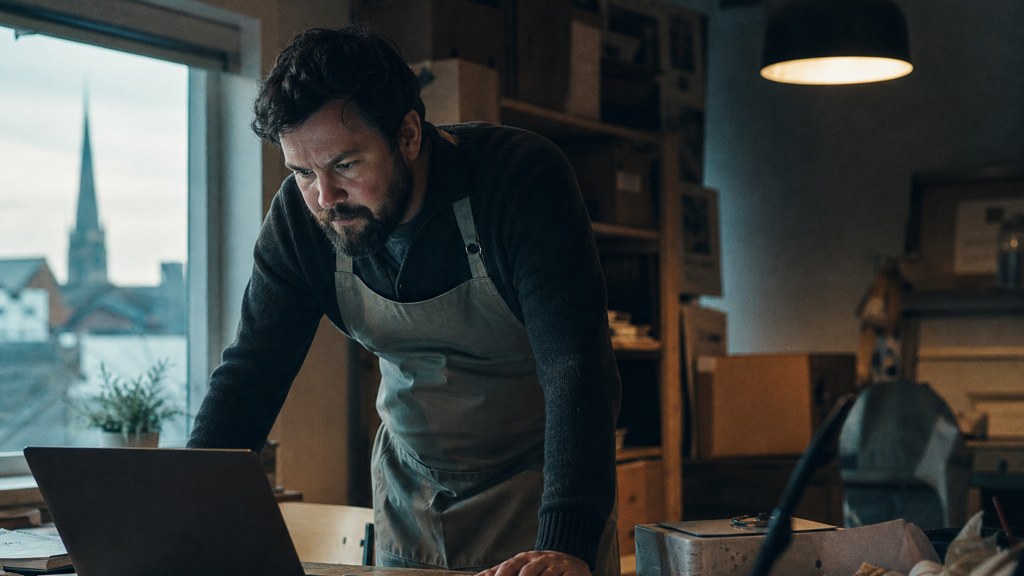 A small-business owner in an apron leaning over a laptop on a cluttered back-office desk late in the day, photographed in Norfolk