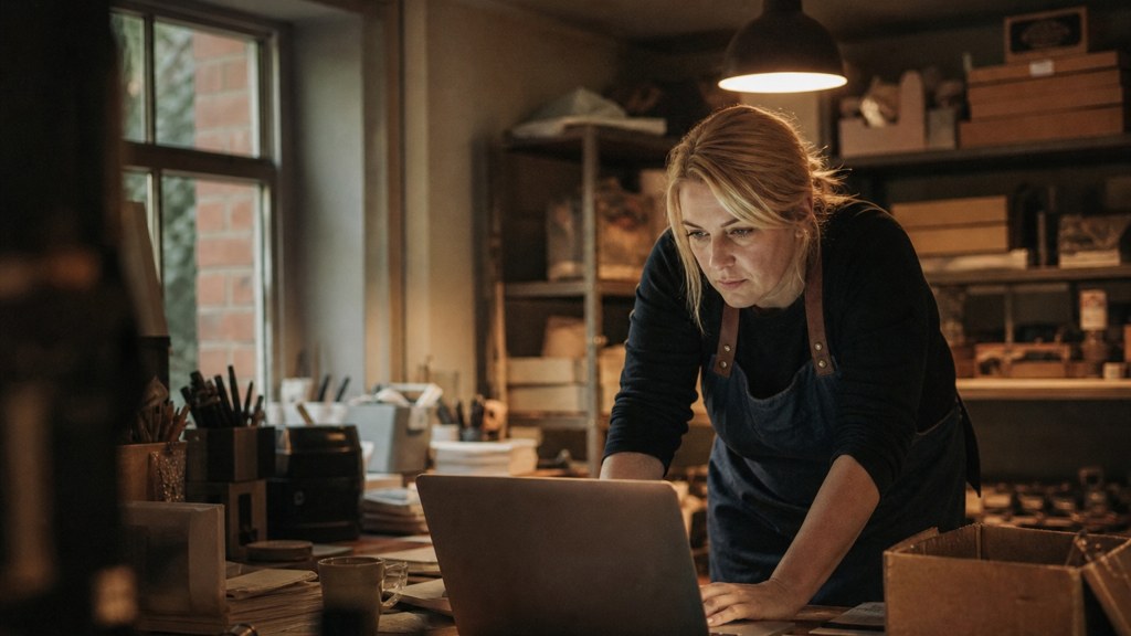 A small-business owner in an apron leaning over a laptop on a cluttered back-office desk late in the day, photographed in Norfolk