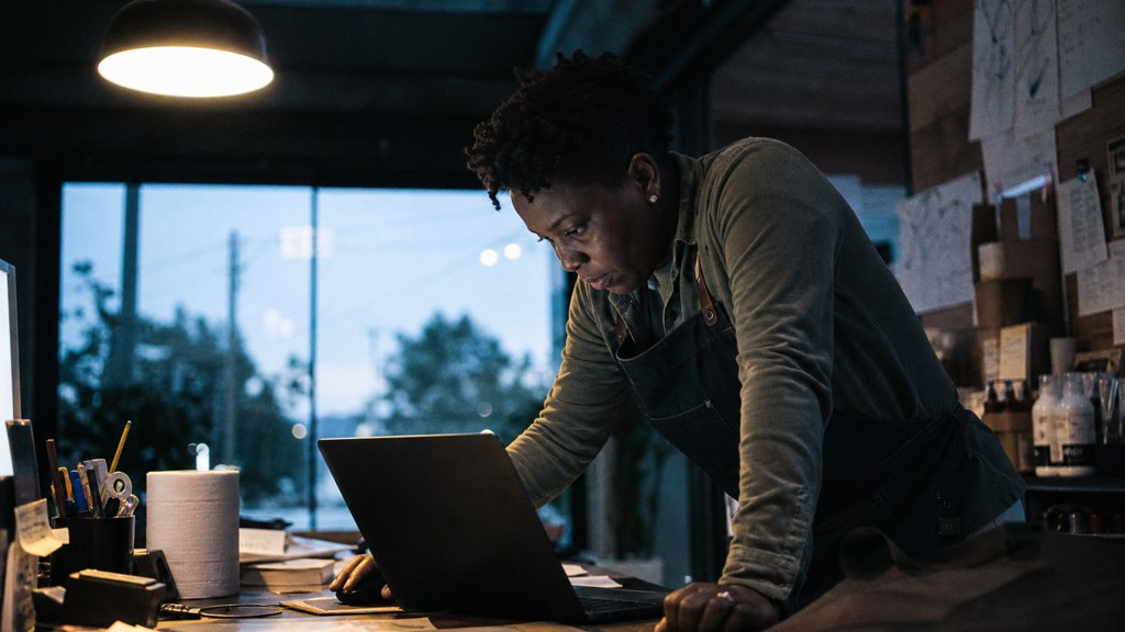 A small-business owner in an apron leaning over a laptop on a cluttered back-office desk late in the day, photographed in Oakland