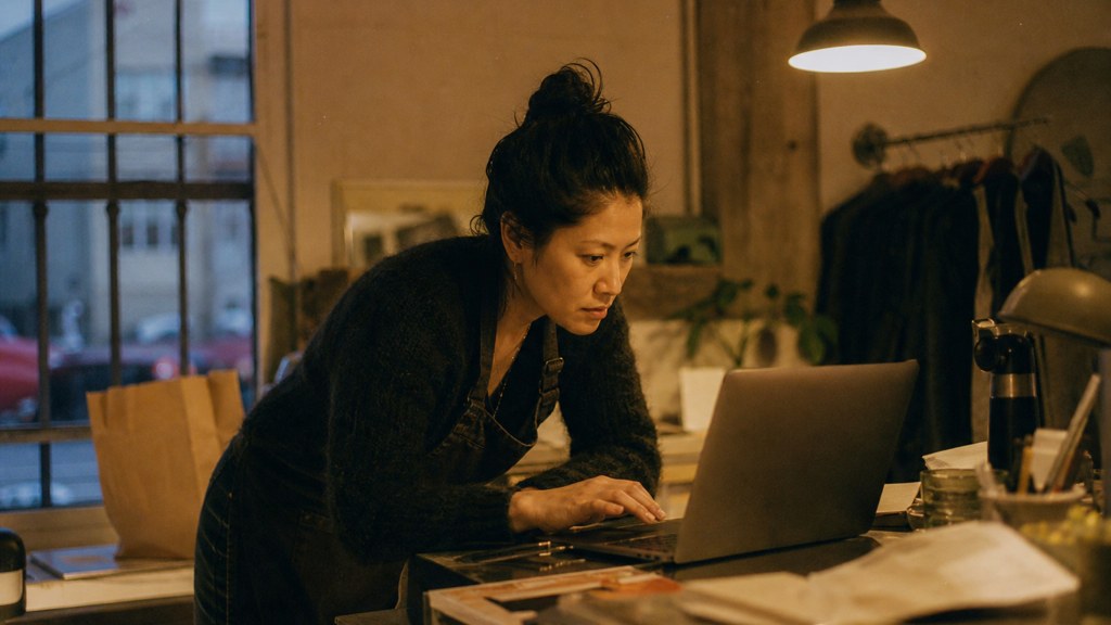 A small-business owner in an apron leaning over a laptop on a cluttered back-office desk late in the day, photographed in Oakland