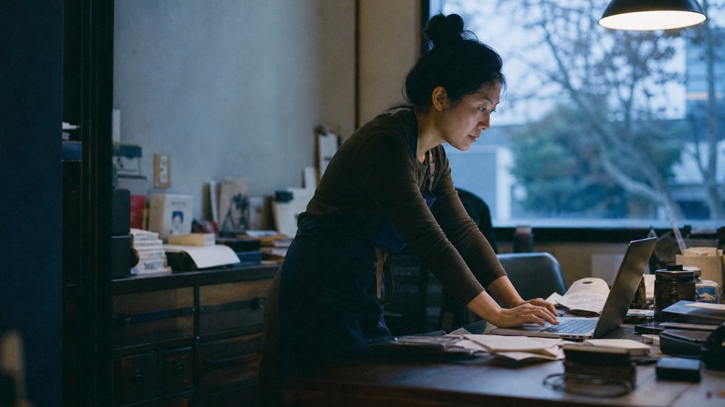 A small-business owner in an apron leaning over a laptop on a cluttered back-office desk late in the day, photographed in Oakland