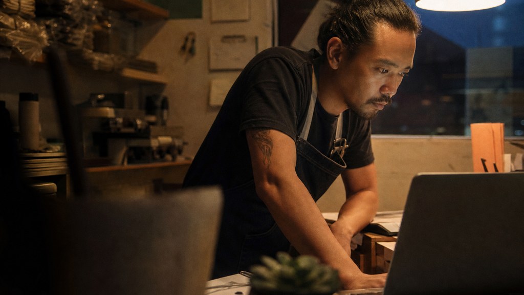A small-business owner in an apron leaning over a laptop on a cluttered back-office desk late in the day, photographed in Oakland