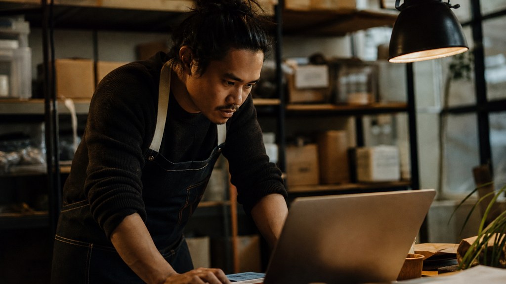 A small-business owner in an apron leaning over a laptop on a cluttered back-office desk late in the day, photographed in Oakland