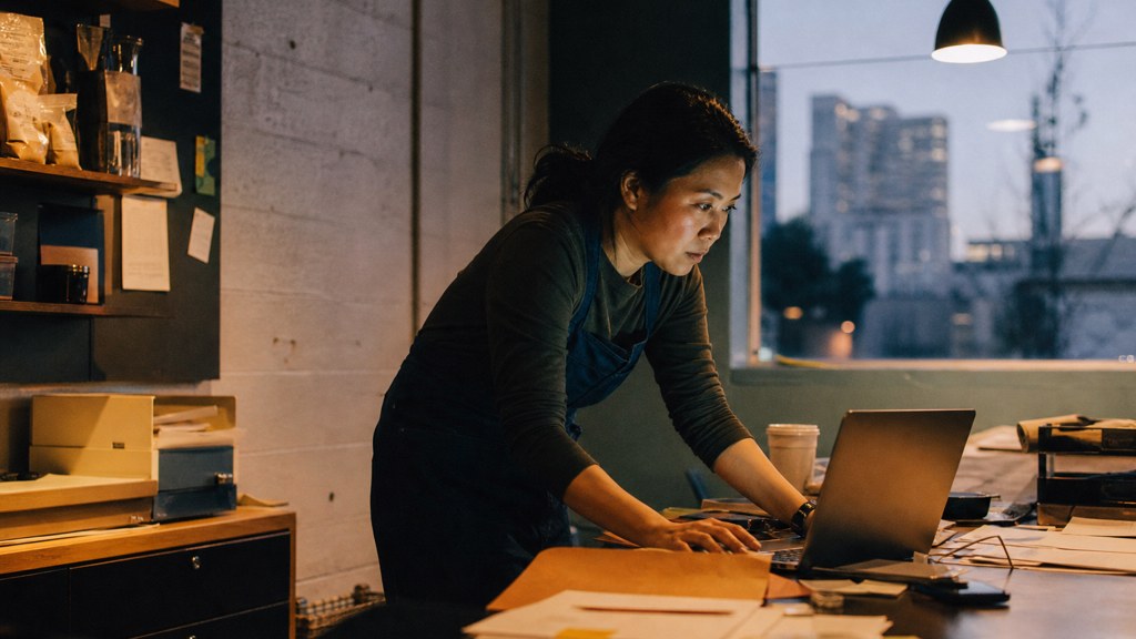 A small-business owner in an apron leaning over a laptop on a cluttered back-office desk late in the day, photographed in Oakland