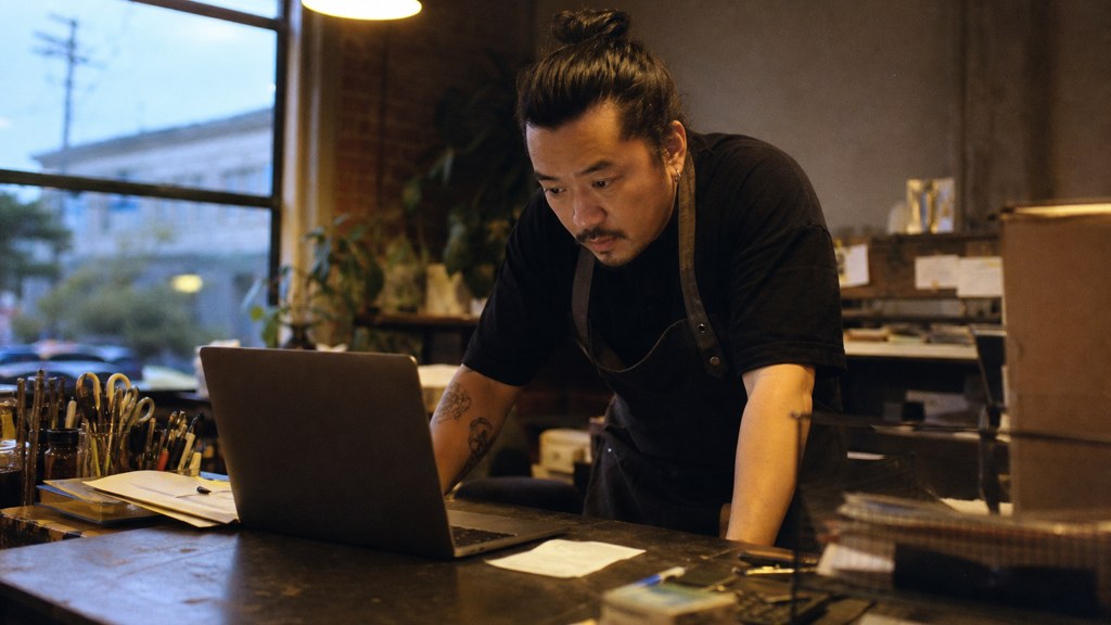 A small-business owner in an apron leaning over a laptop on a cluttered back-office desk late in the day, photographed in Oakland